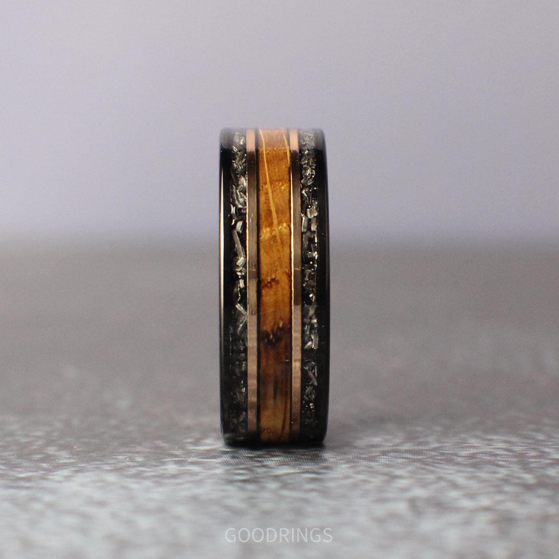 Sophisticated black tungsten wedding band with dual meteorite inlays on the edges, a center strip of deep, charred whiskey barrel wood, and two gleaming rose gold lines, blending strength, history, and elegance in one striking piece.