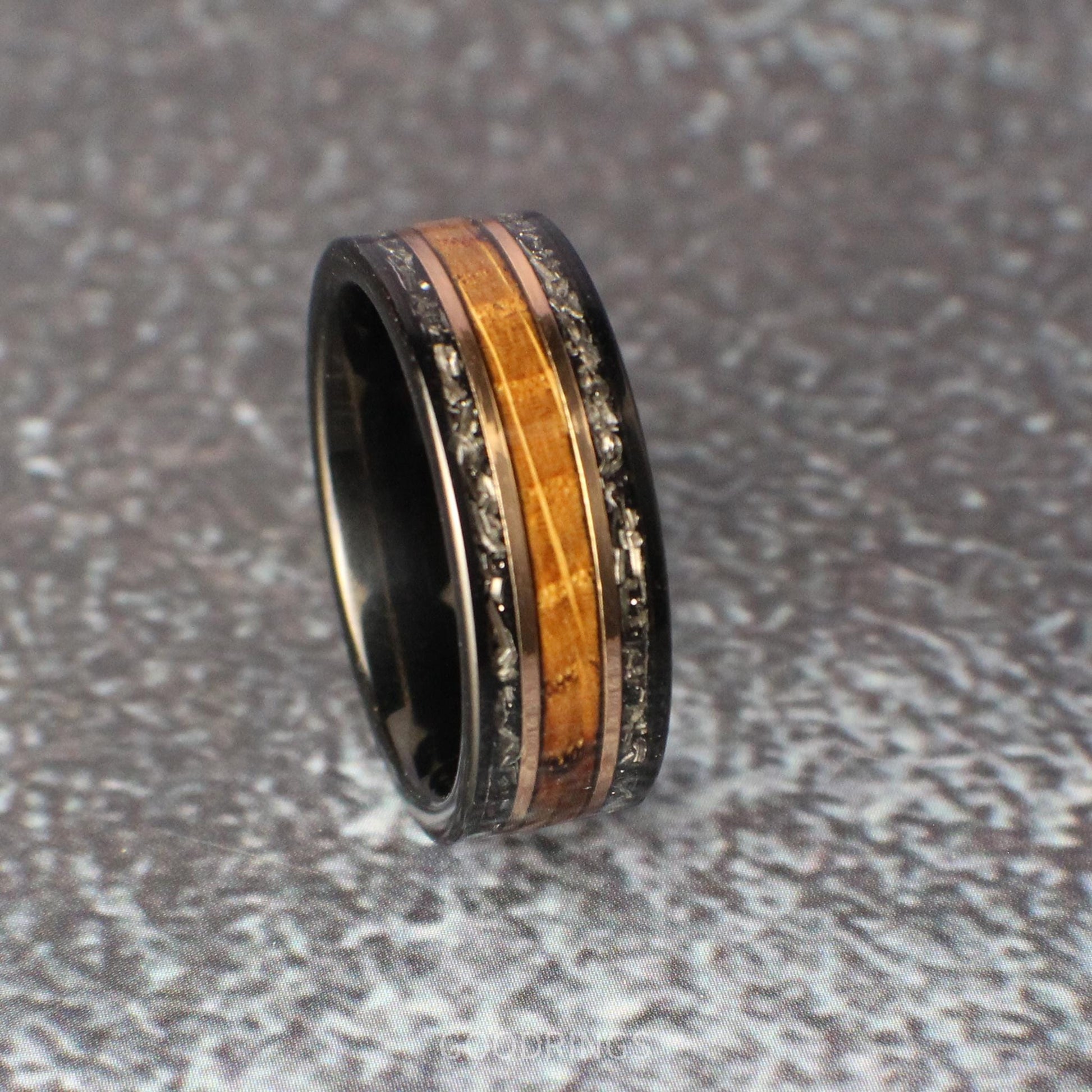 Men’s wedding ring in black tungsten, showcasing two outer bands of meteorite, a center inlay of textured burnt whiskey wood, and rose gold lines separating each element, creating a modern and timeless design.