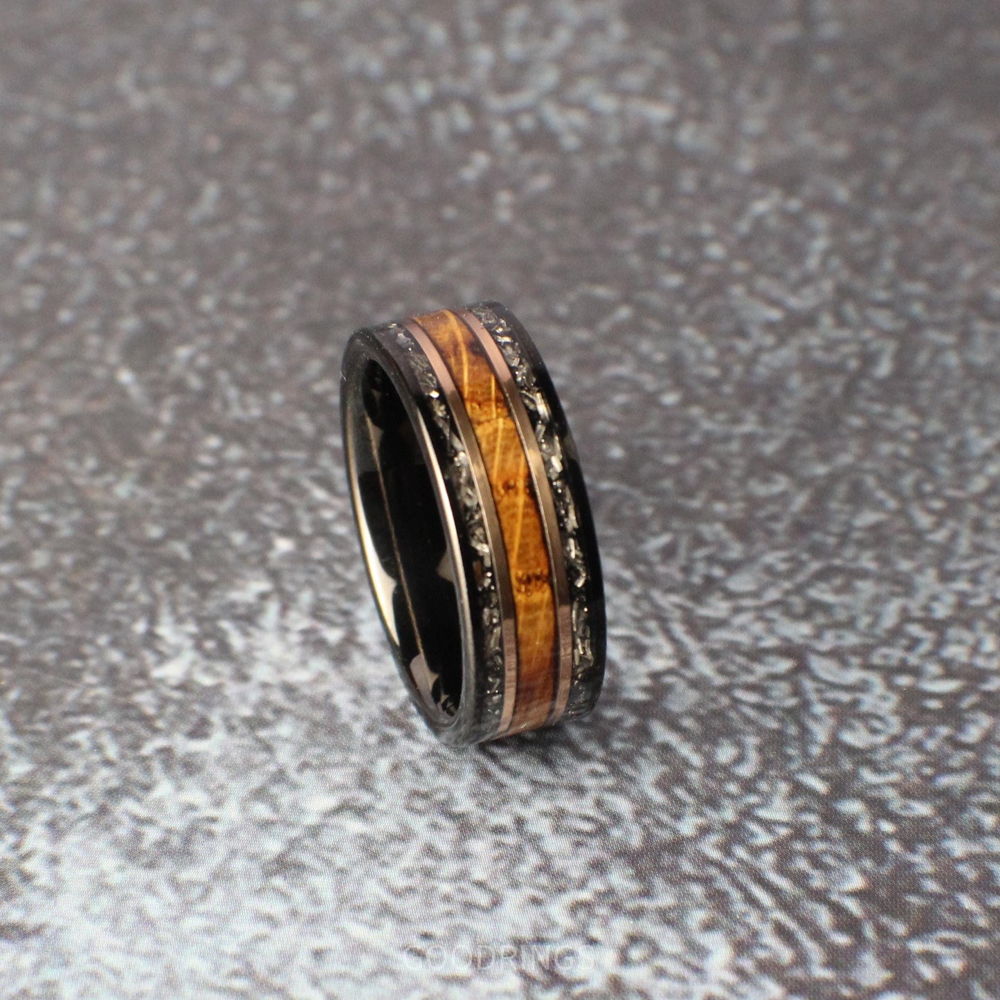 Close-up of a black tungsten men’s ring, featuring contrasting materials: meteorite with subtle patterns on the edges, a charred whiskey wood center, and delicate rose gold dividers, blending contemporary and rustic elements.