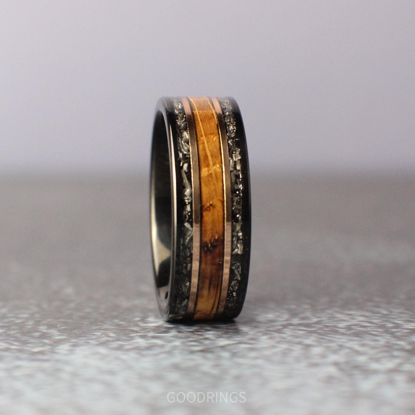 Close-up of a bold black tungsten men’s wedding band, featuring two outer channels of meteorite with intricate, natural patterns, a central inlay of rich burnt whiskey wood, and two sleek rose gold accents separating the materials.