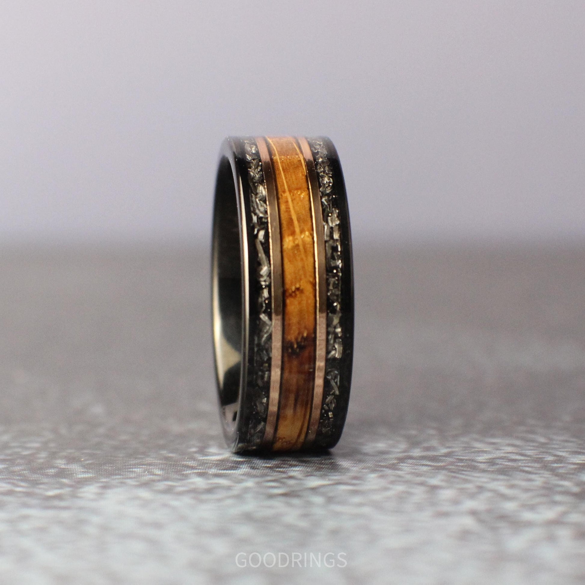 Close-up of a bold black tungsten men’s wedding band, featuring two outer channels of meteorite with intricate, natural patterns, a central inlay of rich burnt whiskey wood, and two sleek rose gold accents separating the materials.